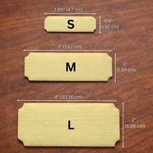 Size comparison of three gold engraved nameplates on a wooden surface labeled S, M, and L, with dimensions shown: Small 1.85" x 0.6" (4.7 x 1.52 cm), Medium 3" x 1" (7.62 x 2.54 cm), and Large 4" x 2" (10.16 x 5.08 cm).