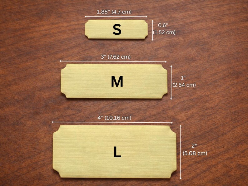 Size comparison of three gold engraved nameplates on a wooden surface labeled S, M, and L, with dimensions shown: Small 1.85" x 0.6" (4.7 x 1.52 cm), Medium 3" x 1" (7.62 x 2.54 cm), and Large 4" x 2" (10.16 x 5.08 cm).