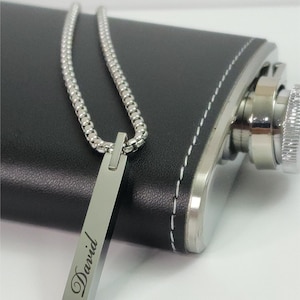 Custom engraved stainless steel bar necklace with the name ‘David,’ shown draped over a black leather flask. Personalized men’s jewelry, custom name necklace, groomsmen gift, anniversary or birthday gift for him.