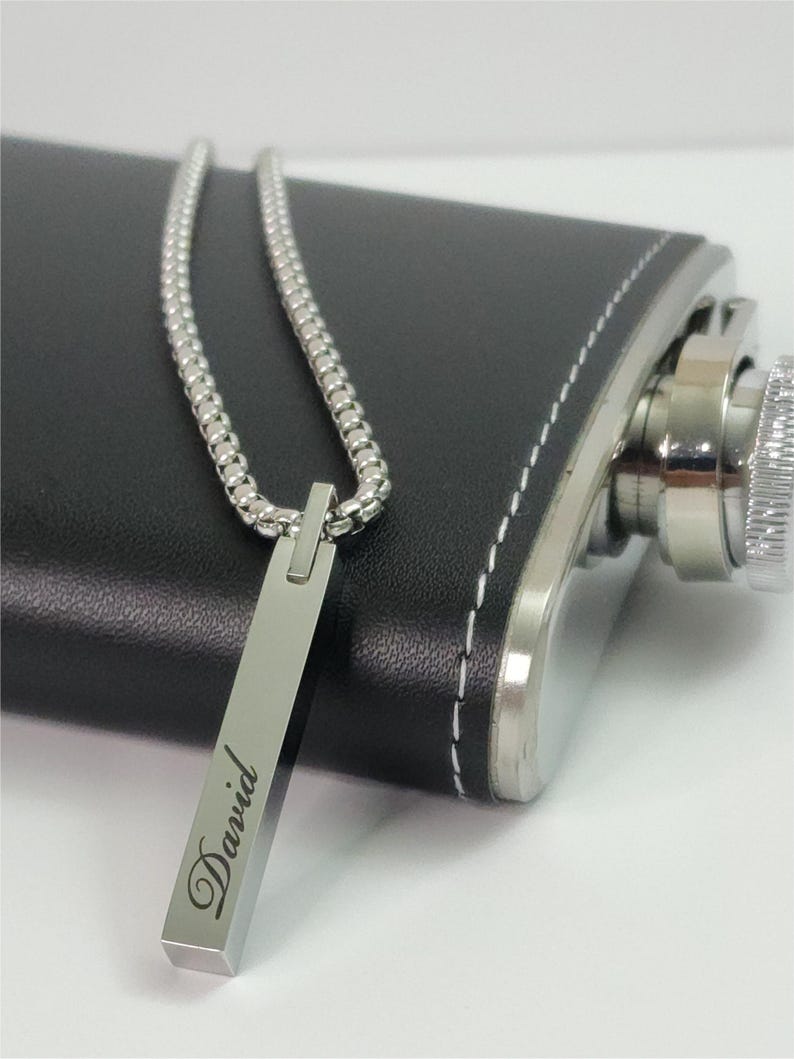 Custom engraved stainless steel bar necklace with the name ‘David,’ shown draped over a black leather flask. Personalized men’s jewelry, custom name necklace, groomsmen gift, anniversary or birthday gift for him.