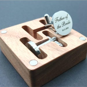 Puede incluir: Una caja de madera con dos gemelos plateados. Un gemelo es redondo con el texto "Father of the Bride" y la fecha "10.11.2025". La caja es de madera marr&oacute;n claro y tiene cierre magn&eacute;tico.