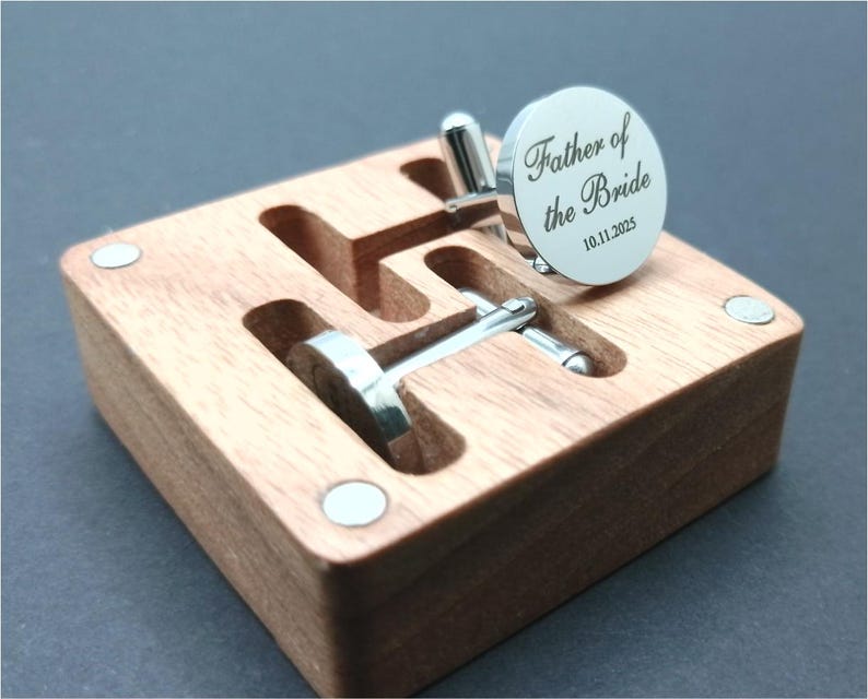 Puede incluir: Una caja de madera con dos gemelos plateados. Un gemelo es redondo con el texto "Father of the Bride" y la fecha "10.11.2025". La caja es de madera marr&oacute;n claro y tiene cierre magn&eacute;tico.
