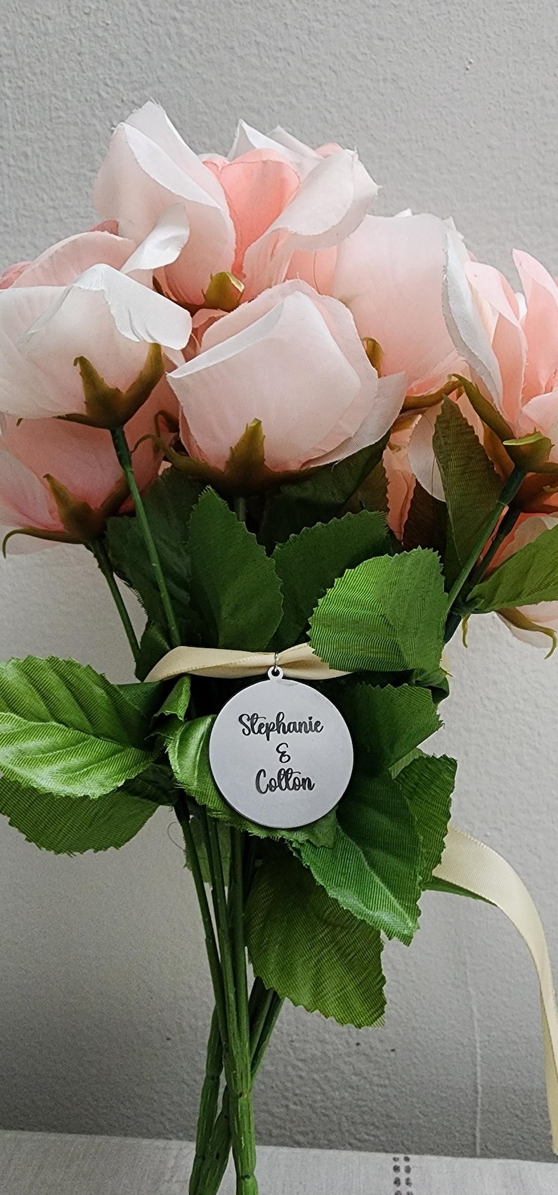 K&ouml;nnte beinhalten: Ein Strau&szlig; aus k&uuml;nstlichen hellrosa Rosen mit gr&uuml;nen Stoffbl&auml;ttern, zusammengebunden mit einem cremefarbenen Band. Ein runder, silberfarbener Metallanh&auml;nger h&auml;ngt an dem Band und tr&auml;gt den schwarzen Gravurtext Stephanie & Colton.