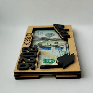 Ultra-low side profile view of a personalized laser-engraved birch wood graduation money holder reading "Andrew – Class of 2026," clearly showing the open bottom bill slot and precise multi-layer wood sandwich construction, with a $100 bill visible inside, raised black laser-cut "2026" numbers and "Class of" text on the left side, a laser-cut graduation cap accent on the right, and a black script name tag on the top edge, photographed against a clean white background.