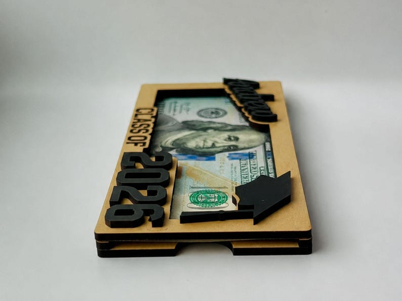 Ultra-low side profile view of a personalized laser-engraved birch wood graduation money holder reading "Andrew – Class of 2026," clearly showing the open bottom bill slot and precise multi-layer wood sandwich construction, with a $100 bill visible inside, raised black laser-cut "2026" numbers and "Class of" text on the left side, a laser-cut graduation cap accent on the right, and a black script name tag on the top edge, photographed against a clean white background.