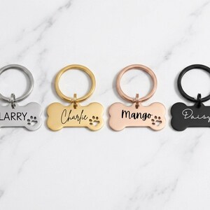 Four custom bone-shaped pet ID tags in silver, gold, rose gold, and black finishes, engraved with the names 'LARRY', 'Charlie', 'Mango', and 'Daisy' in different font styles. Each tag features a matching split key ring and paw print cutout, displayed on a white marble background.