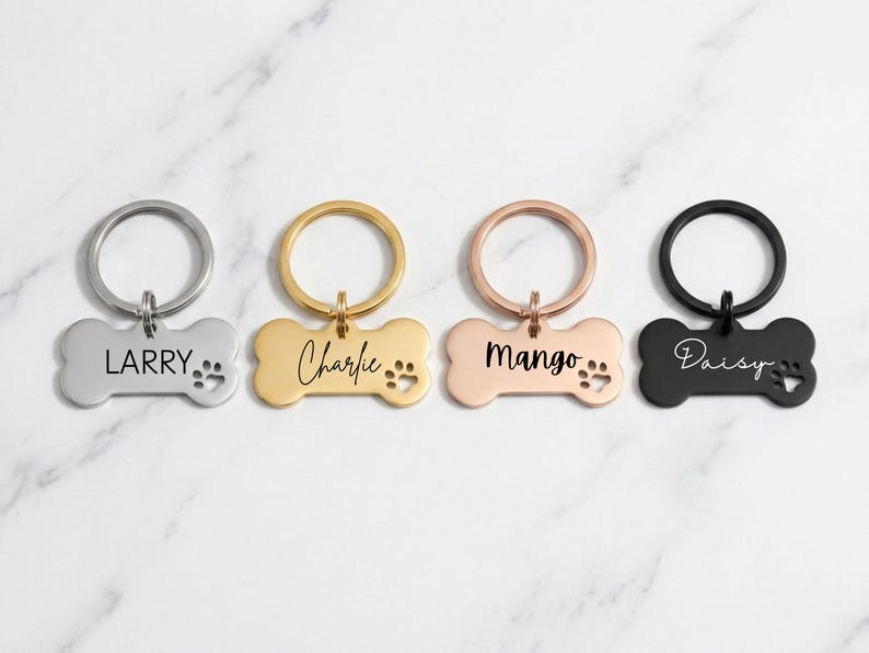 Four custom bone-shaped pet ID tags in silver, gold, rose gold, and black finishes, engraved with the names 'LARRY', 'Charlie', 'Mango', and 'Daisy' in different font styles. Each tag features a matching split key ring and paw print cutout, displayed on a white marble background.