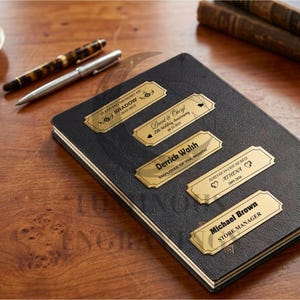 Five engraved gold metal nameplates displayed on a black leather notebook placed on a wooden desk, including memorial, anniversary, employee, and manager titles; a cup of tea, two pens, and stacked books sit in the background, creating a warm workspace setting.