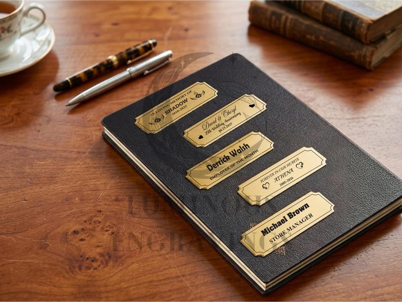 Five engraved gold metal nameplates displayed on a black leather notebook placed on a wooden desk, including memorial, anniversary, employee, and manager titles; a cup of tea, two pens, and stacked books sit in the background, creating a warm workspace setting.