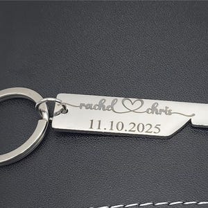 A silver-colored keychain with a rectangular tag and a bottle opener design. The tag is engraved with the names "rachel" and "chris", a heart symbol, and the date "11.10.2025". The keychain is on a dark surface.