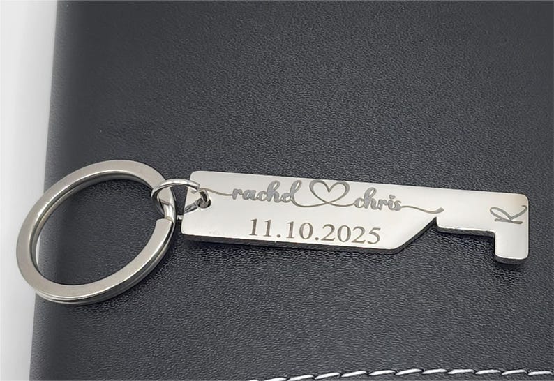 A silver-colored keychain with a rectangular tag and a bottle opener design. The tag is engraved with the names "rachel" and "chris", a heart symbol, and the date "11.10.2025". The keychain is on a dark surface.