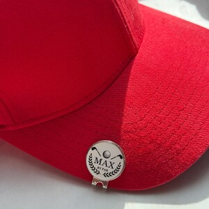 Puede incluir: Una gorra de b&eacute;isbol de color rojo brillante con un marcador de pelota de golf circular de metal plateado sujeto a la visera. El marcador muestra un gr&aacute;fico negro de dos palos de golf cruzados, una pelota y el texto MAX BY PAR con laureles.
