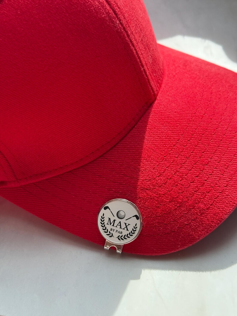 Puede incluir: Una gorra de b&eacute;isbol de color rojo brillante con un marcador de pelota de golf circular de metal plateado sujeto a la visera. El marcador muestra un gr&aacute;fico negro de dos palos de golf cruzados, una pelota y el texto MAX BY PAR con laureles.