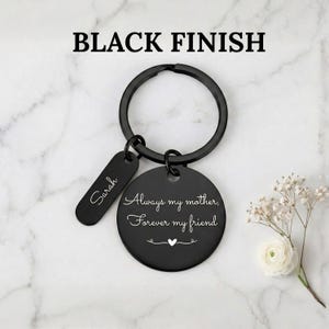 Black finish personalized keychain for mom with engraved message “Always my mother, forever my friend” and custom name tag, displayed on a marble background with delicate white flowers.