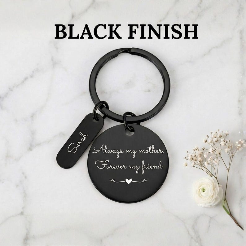 Black finish personalized keychain for mom with engraved message “Always my mother, forever my friend” and custom name tag, displayed on a marble background with delicate white flowers.