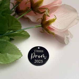 May include: A black circular tag with the text "CHHS Prom 2025" in white script. The tag is placed near pink artificial roses and green leaves.