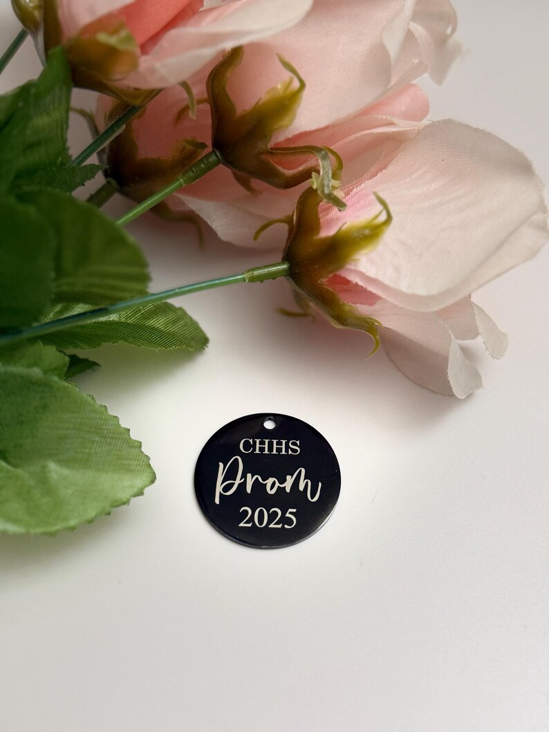 May include: A black circular tag with the text "CHHS Prom 2025" in white script. The tag is placed near pink artificial roses and green leaves.