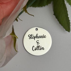 May include: A round, silver-colored charm with the names "Stephanie & Colton" in black script. The charm has a small hole at the top for hanging. The background includes pink and green floral elements.