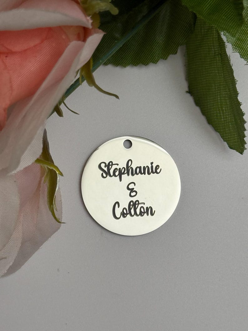 May include: A round, silver-colored charm with the names "Stephanie & Colton" in black script. The charm has a small hole at the top for hanging. The background includes pink and green floral elements.