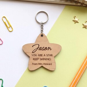 Close-up flat lay of a personalized wooden star-shaped keychain engraved with the name "Jason" and the message, "YOU ARE A STAR KEEP SHINING! From Mrs. Howard." The laser-cut keychain is attached to a silver metal chain and displayed on a modern white and pastel green background with stationery accents, highlighting it as a thoughtful teacher appreciation or student gift.