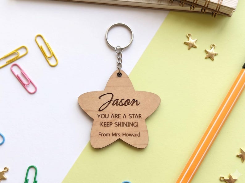 Close-up flat lay of a personalized wooden star-shaped keychain engraved with the name "Jason" and the message, "YOU ARE A STAR KEEP SHINING! From Mrs. Howard." The laser-cut keychain is attached to a silver metal chain and displayed on a modern white and pastel green background with stationery accents, highlighting it as a thoughtful teacher appreciation or student gift.