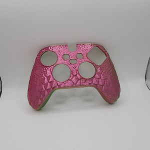 Peut inclure: Coque de manette rose et verte irisée avec un motif texturé et écailleux. La coque comporte des découpes pour les boutons et les joysticks et est conçue pour s'adapter à une manette de jeu. La coque est en plastique.