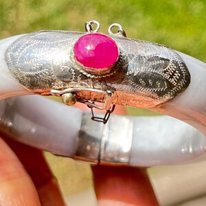 May include: A hinged bangle bracelet featuring a smooth, pale lavender-grey stone band. The center section is crafted from etched silver metal and is accented with a single, vibrant, oval-shaped hot pink cabochon stone set in a silver bezel.