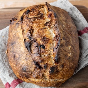 Può includere: Una pagnotta di pane artigianale appena sfornata con una crosta dorata e macchie scure e caramellate. Il pane è adagiato su un panno di lino grigio con una striscia rossa, posto su un tagliere di legno. La consistenza del pane è ruvida e rustica.