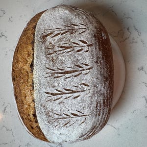 May include: A rustic loaf of bread with a dark brown crust and a white, flour-dusted top. The top is decorated with leaf-like patterns. The bread is placed on a white plate, with a light-coloured countertop in the background.