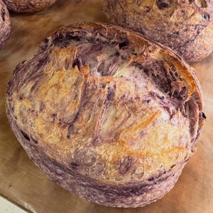May include: A round loaf of sourdough bread with a deep purple-tinted crust and a golden-brown, scored top. The bread features a rustic, uneven texture with visible air pockets and a dark, flour-dusted exterior, showcasing a baked artisan loaf.