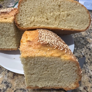 May include: A loaf of golden-brown bread cut into large pieces, featuring a porous, light-colored interior crumb and a crust topped with a generous sprinkling of small, white sesame seeds, presented on a white plate for serving.