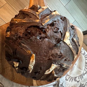 May include: A loaf of dark brown bread with a textured crust, resting on a wooden cutting board. The bread has light-colored accents and appears freshly baked. The wooden board has a handle.