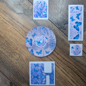May include: A selection of decorative stickers with a butterfly design. The stickers display blue butterflies, floral patterns, and botanical elements on a lilac background. The stickers are in various shapes and sizes.