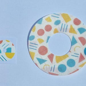 May include: Two round stickers with a white background and a geometric pattern in blue, yellow, and pink. The smaller sticker is approximately 5 cm in diameter and the larger sticker is approximately 10 cm in diameter.