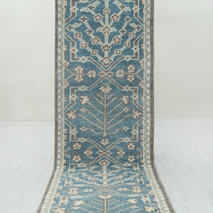 May include: A rectangular, blue runner rug with a detailed geometric pattern. The rug features a central design with stylized floral and tree-like motifs in shades of beige and brown. The border is a darker brown, and the rug is displayed on a light-colored surface.