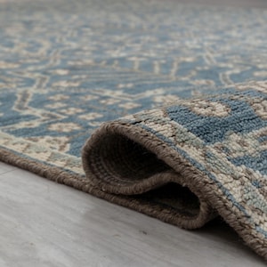 May include: A rolled-up area rug with a detailed pattern in shades of blue, beige, and gray. The rug's edge is a darker brown, and the texture is visible. The rug is on a light gray surface.