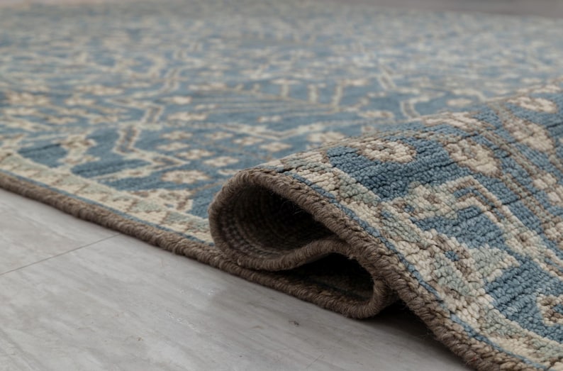 May include: A rolled-up area rug with a detailed pattern in shades of blue, beige, and gray. The rug's edge is a darker brown, and the texture is visible. The rug is on a light gray surface.