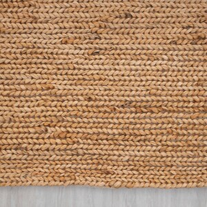 May include: Close-up of a rectangular, woven jute rug. The rug has a natural, light brown color and a thick, braided texture. The rug is placed on a light gray surface.