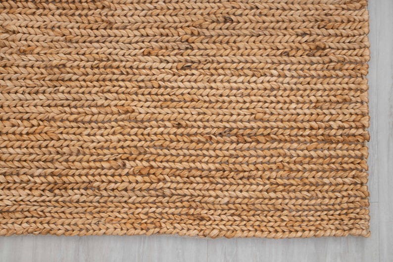 May include: Close-up of a rectangular, woven jute rug. The rug has a natural, light brown color and a thick, braided texture. The rug is placed on a light gray surface.