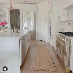 Puede incluir: Una cocina moderna y luminosa con gabinetes blancos, electrodomésticos de acero inoxidable y pisos de madera clara. Una alfombra corredora tejida con borde festoneado recorre el centro. Los herrajes y accesorios dorados añaden un toque de elegancia.