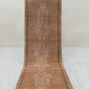 Puede incluir: Una alfombra de pasillo rectangular marrón con un detallado patrón geométrico en color beige claro. La alfombra tiene flecos en el borde superior y un borde marrón más oscuro. El diseño presenta formas y líneas repetidas.