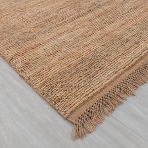 Puede incluir: Una alfombra de yute de tono natural con un diseño tejido texturizado. La alfombra presenta un borde con flecos y una paleta de colores neutros, adecuada para varios estilos de interior. Se ve una esquina de la alfombra.