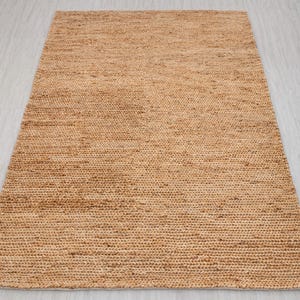 May include: A rectangular, woven jute rug in a natural tan color. The rug has a textured surface with visible fibers and a simple, minimalist design. This rug is suitable for various interior decor styles.