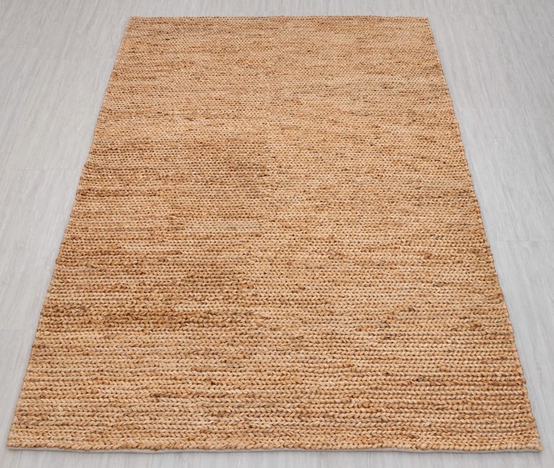 May include: A rectangular, woven jute rug in a natural tan color. The rug has a textured surface with visible fibers and a simple, minimalist design. This rug is suitable for various interior decor styles.