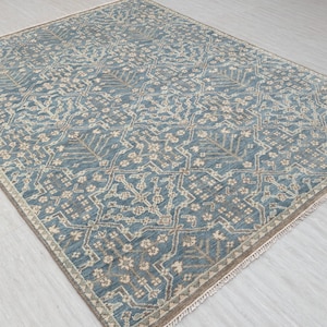 Blue flat-weave area rug with an intricate geometric and floral pattern in beige and cream tones, laid out on a light wood floor and photographed at an angle.