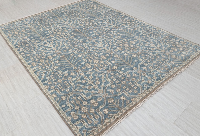 Blue flat-weave area rug with an intricate geometric and floral pattern in beige and cream tones, laid out on a light wood floor and photographed at an angle.