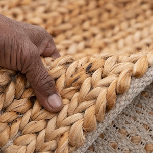 May include: Close-up of a natural fiber rug with a braided border. The rug is made of light brown jute fibers, with a beige backing. The texture of the rug is visible, showing the woven pattern. The rug is partially folded over.