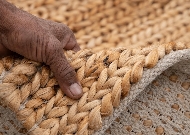 May include: Close-up of a natural fiber rug with a braided border. The rug is made of light brown jute fibers, with a beige backing. The texture of the rug is visible, showing the woven pattern. The rug is partially folded over.