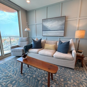 Puede incluir: Salón con un sofá claro, cojines azules y una mesa de centro de madera. Una alfombra azul estampada cubre el suelo. Una pintura de un paisaje marino cuelga de la pared. También se ven un sillón gris y una lámpara de pie.