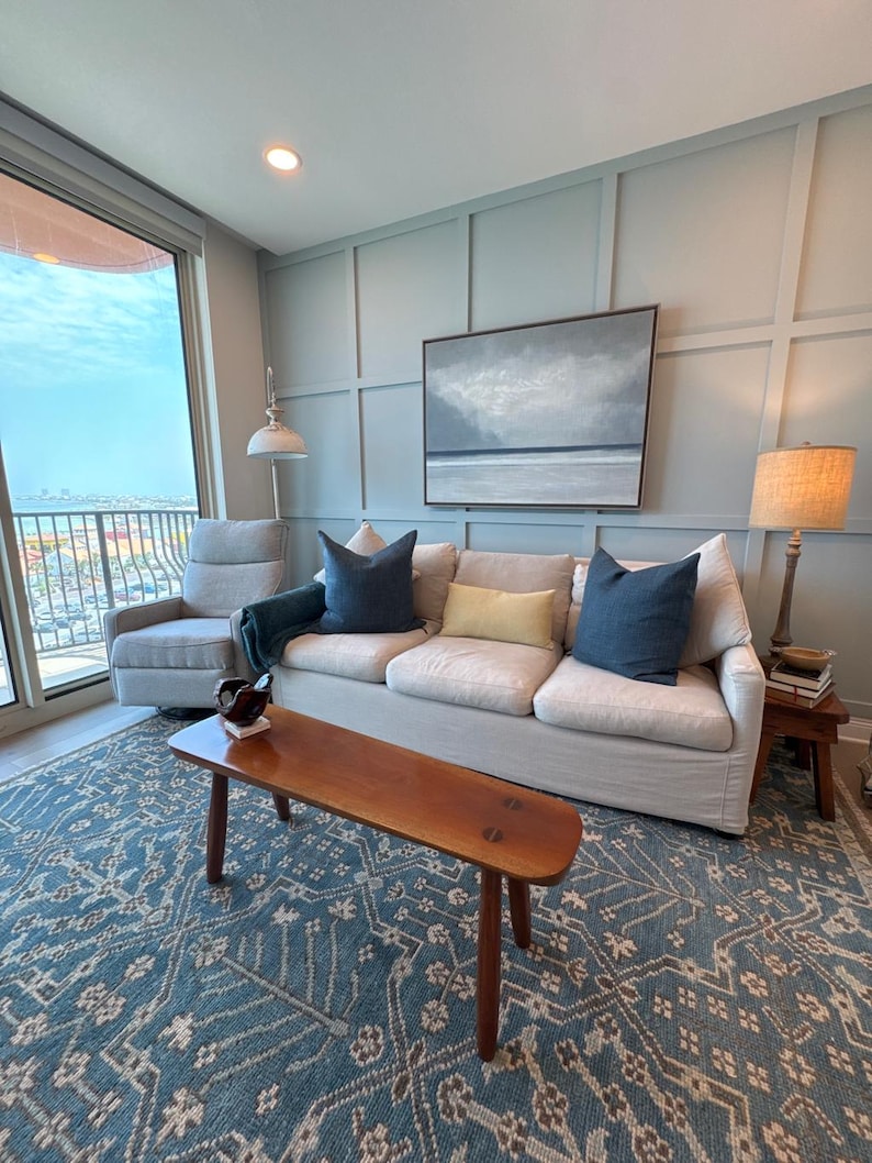 May include: A living room with a light-colored sofa, blue throw pillows, and a wooden coffee table. A blue patterned rug covers the floor. A painting of a seascape hangs on the wall. A gray armchair and a floor lamp are also visible.